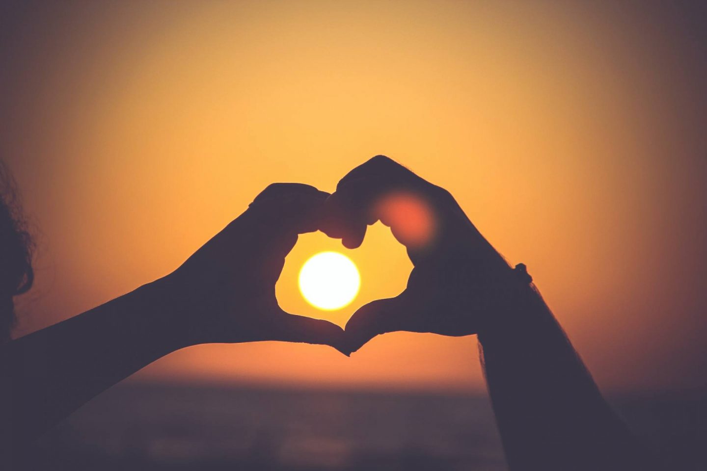 wo people hold their hands up in front of a sunset, making a "heart" shape.